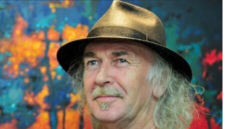 Man met hoed voor kleurrijk schilderij met bomen en huizen op de achtergrond.