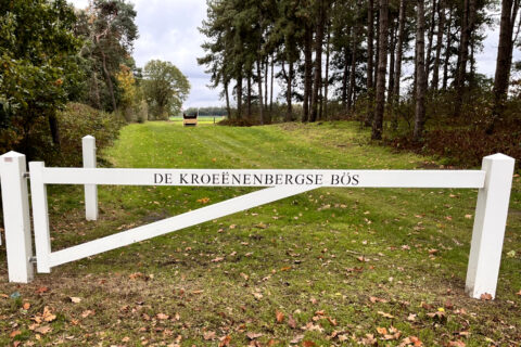 Witte poort met opschrift "De Kroënenbergse Bös" bij groen bospad en bomenrij.