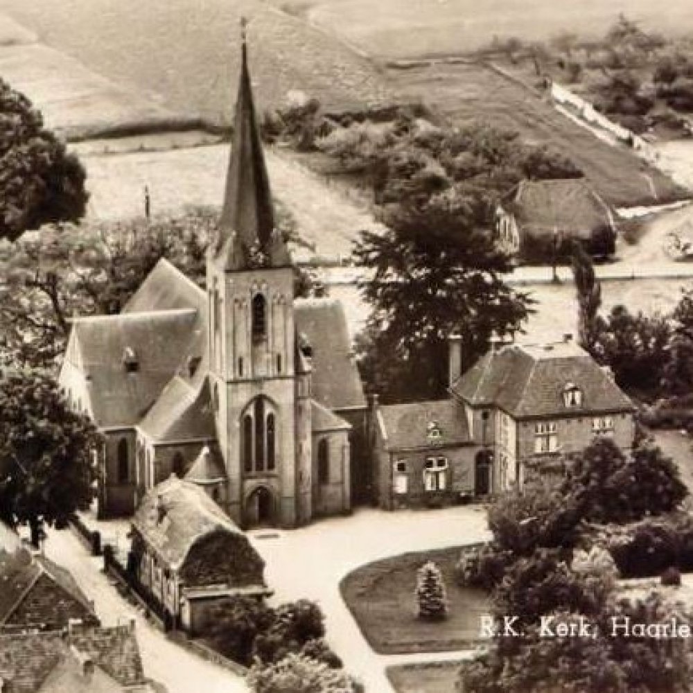 Luchtfoto van een oude kerk met hoge torenspits omgeven door bomen en landelijke gebouwen.