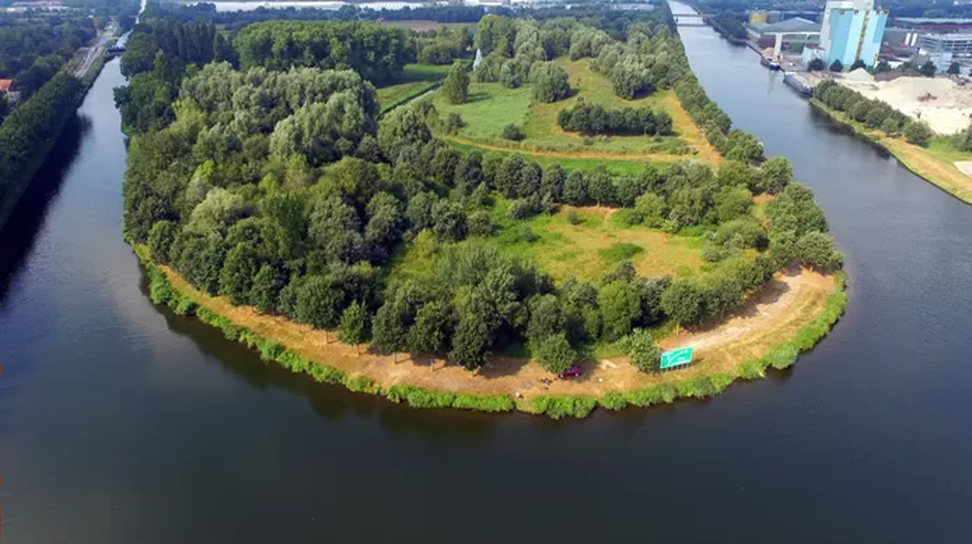 Luchtfoto van een groen bosrijk schiereiland omringd door een rivier en industrie op de achtergrond.