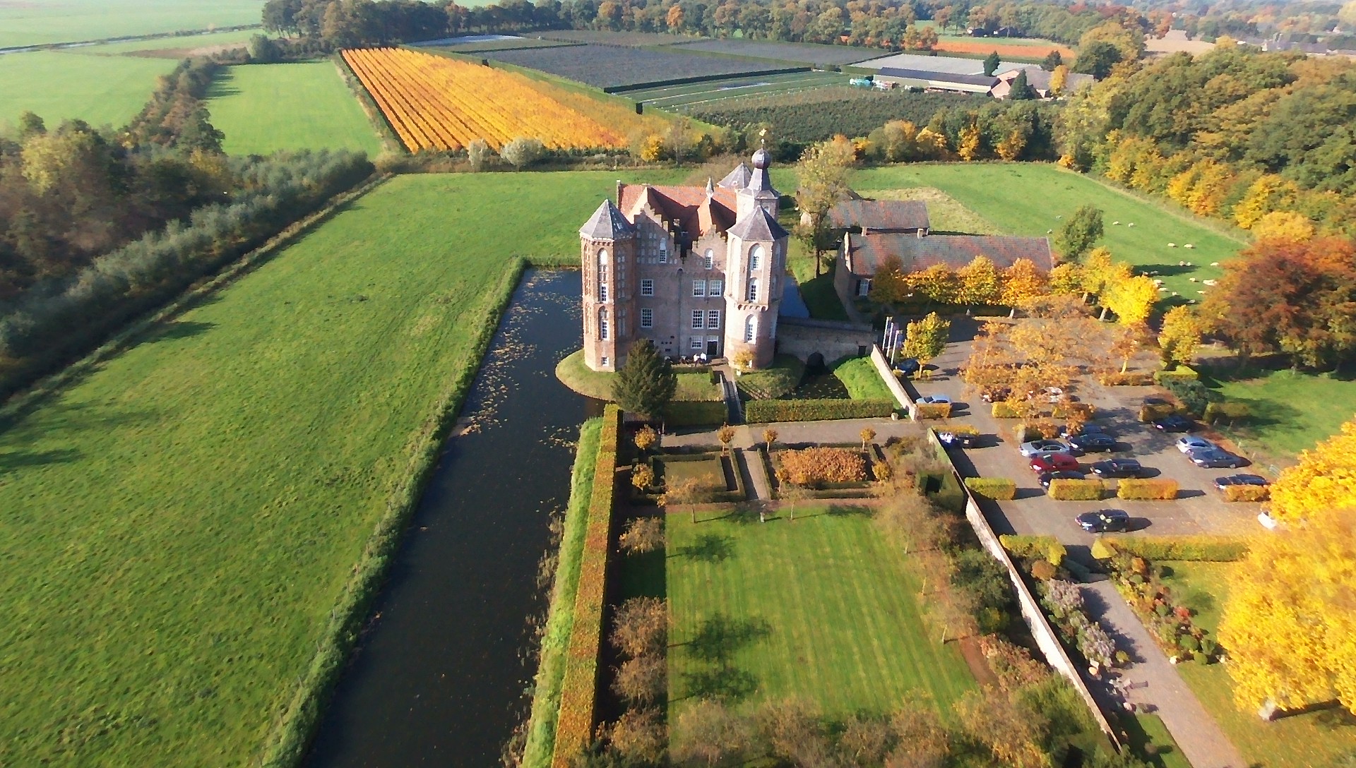 Luchtfoto van een kasteel met een slotgracht, omgeven door velden en een parkeerplaats.