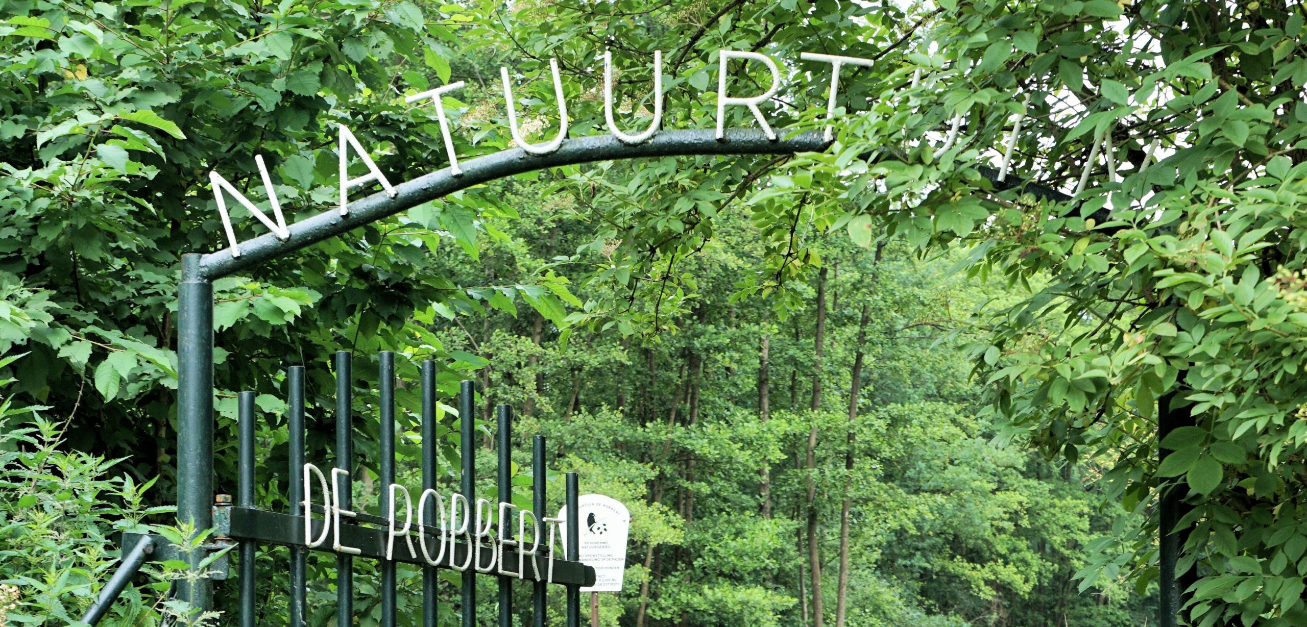 Toegangspoort met "Natuur" en "De Robbert", omgeven door dichte groene bomen en struiken.