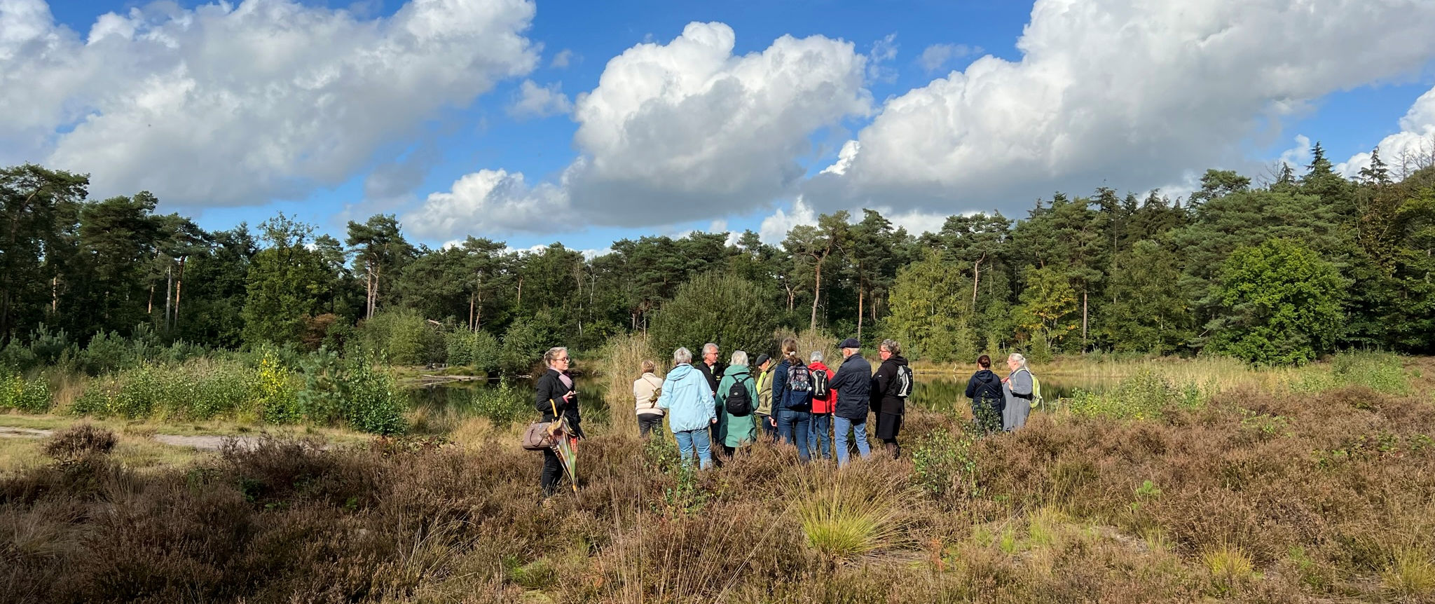 Groep mensen wandelt in een bosrijke omgeving onder een blauwe lucht met wolken.