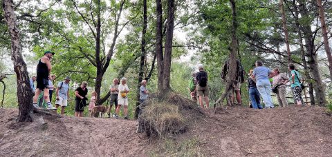 Groep mensen met een hond in een bosrijke omgeving, omringd door bomen op een lichte helling.