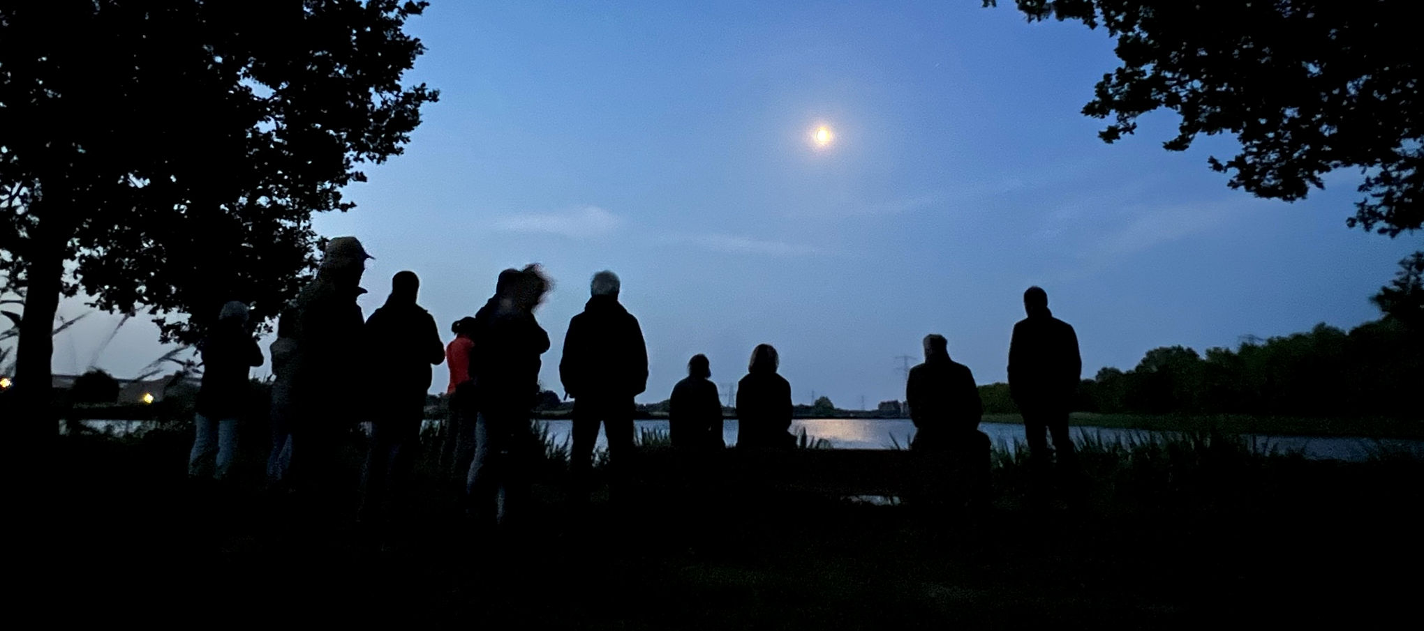 Silhouetten van mensen bij een meer, met de maan verlicht in de avondlucht.