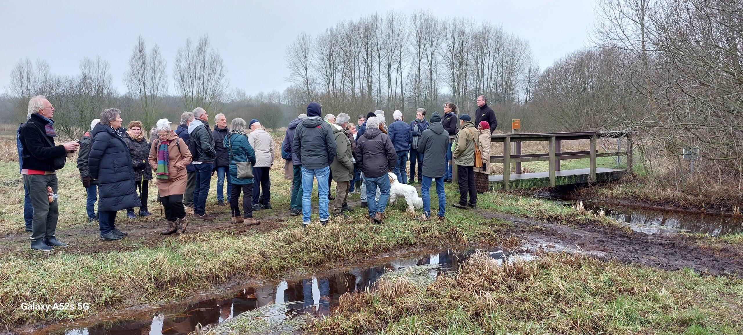 Groep mensen verzameld op een pad naast een houten brug in een bosrijke omgeving.