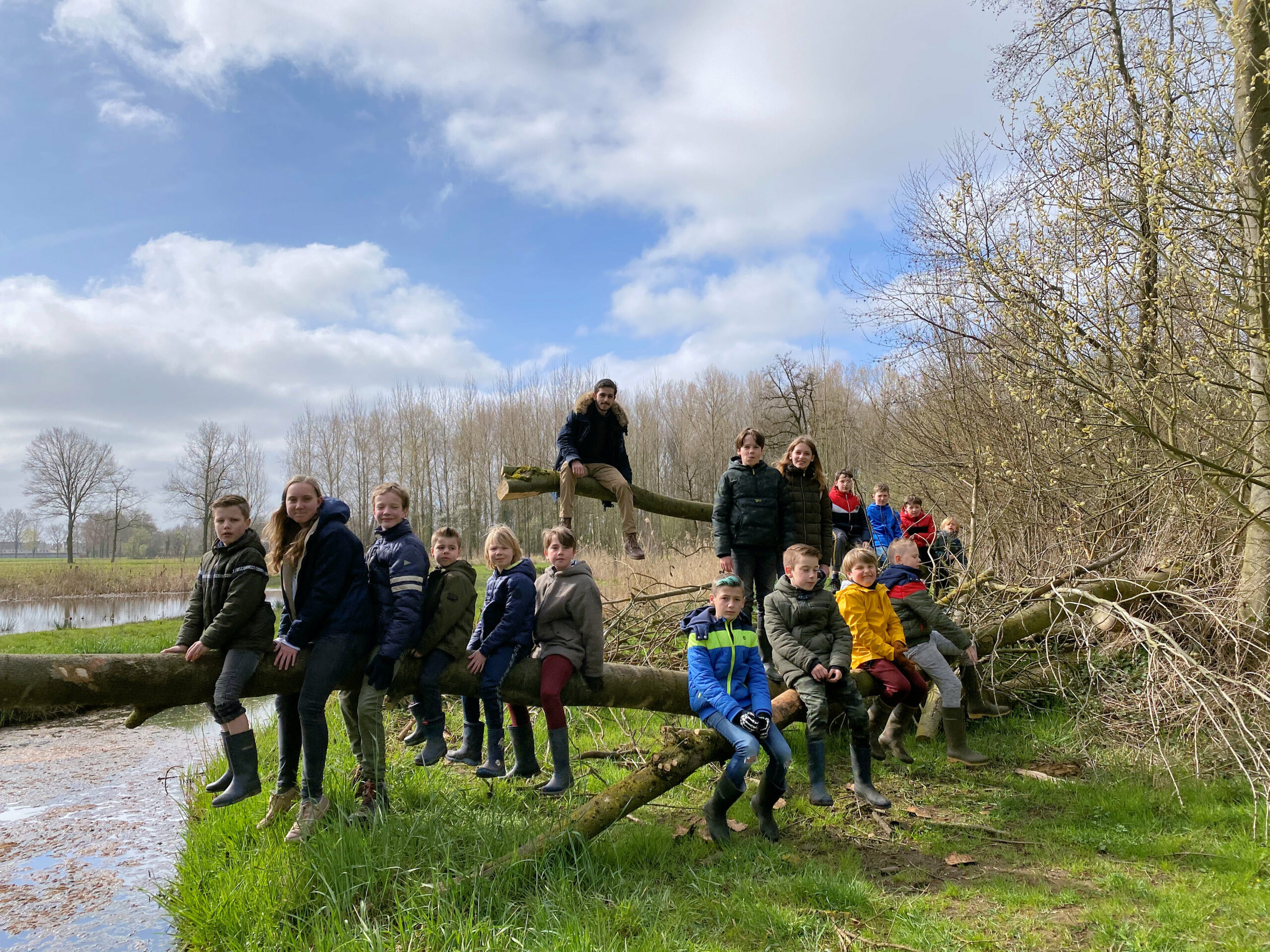 Groep kinderen en een volwassene zitten op een omgevallen boom in een natuurgebied bij een vijver.