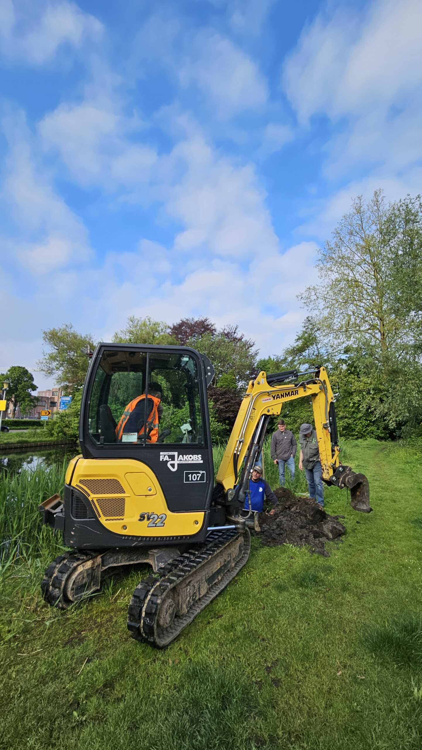 Graafmachine en werkers in groen park onder blauwe lucht met wolken.