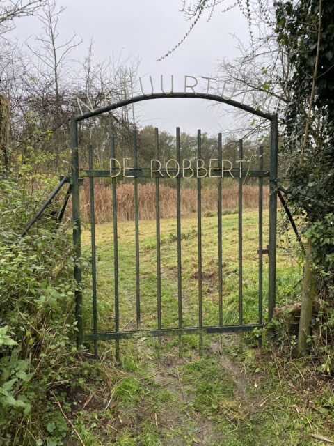 IJzeren poort met tekst "Natuurtuin De Robbert", omgeven door groene planten en begroeid pad.