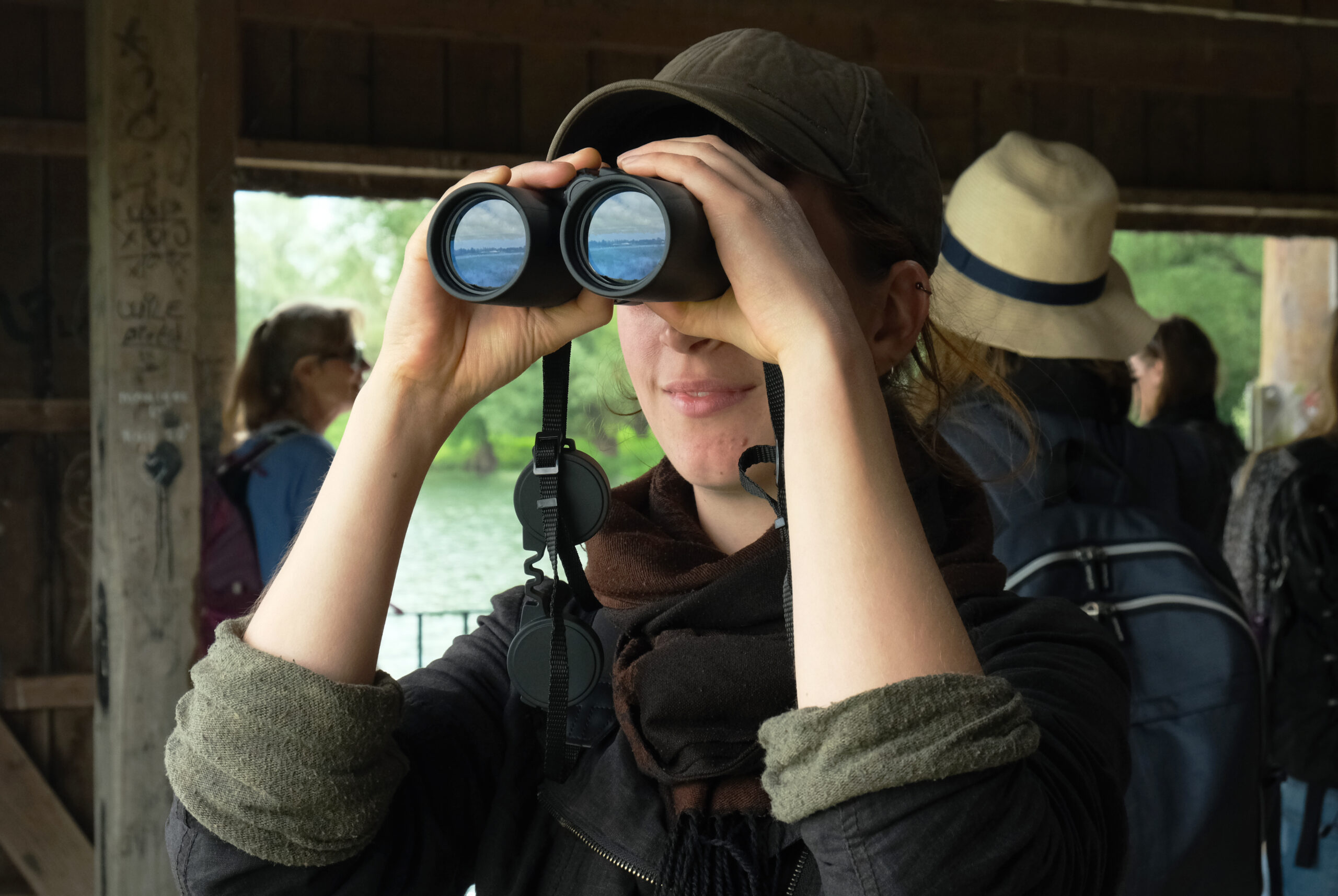 Persoon met pet kijkt door verrekijker; mensen en natuur op achtergrond.