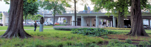 Park met grote bomen, een voetganger, en een kunstwerk; viaduct en auto's op de achtergrond.