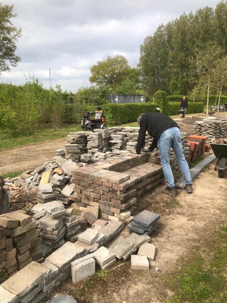 Man stapelt bakstenen op een bouwlocatie buiten, met groen en bomen op de achtergrond.