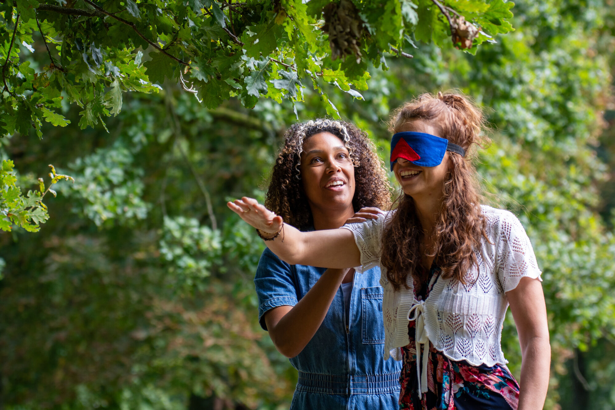 Twee vrouwen in een bos; één geblinddoekt, geleid door de ander, beiden lachend.