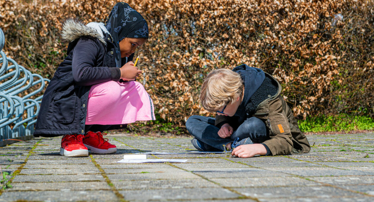 Twee kinderen tekenen op de stoep, omgeven door een heg en fietsenrek.