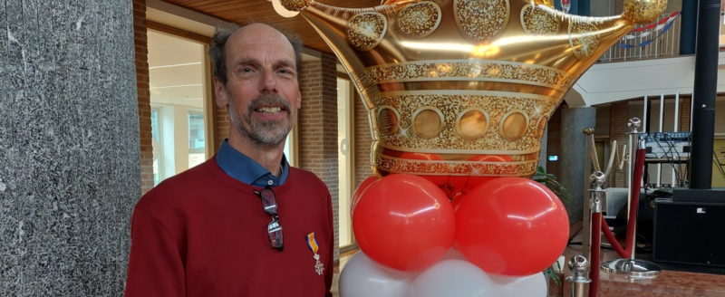 Man met medaille voor gouden kroonballon en rood-witte ballondecoratie binnengebouw.