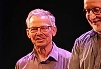 Twee oudere mannen glimlachen op een podium, gekleed in overhemden, tegen een donkere achtergrond.