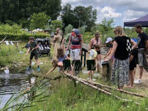 Een groep mensen kijkt toe bij kinderen die met netten aan de oever van een vijver spelen.