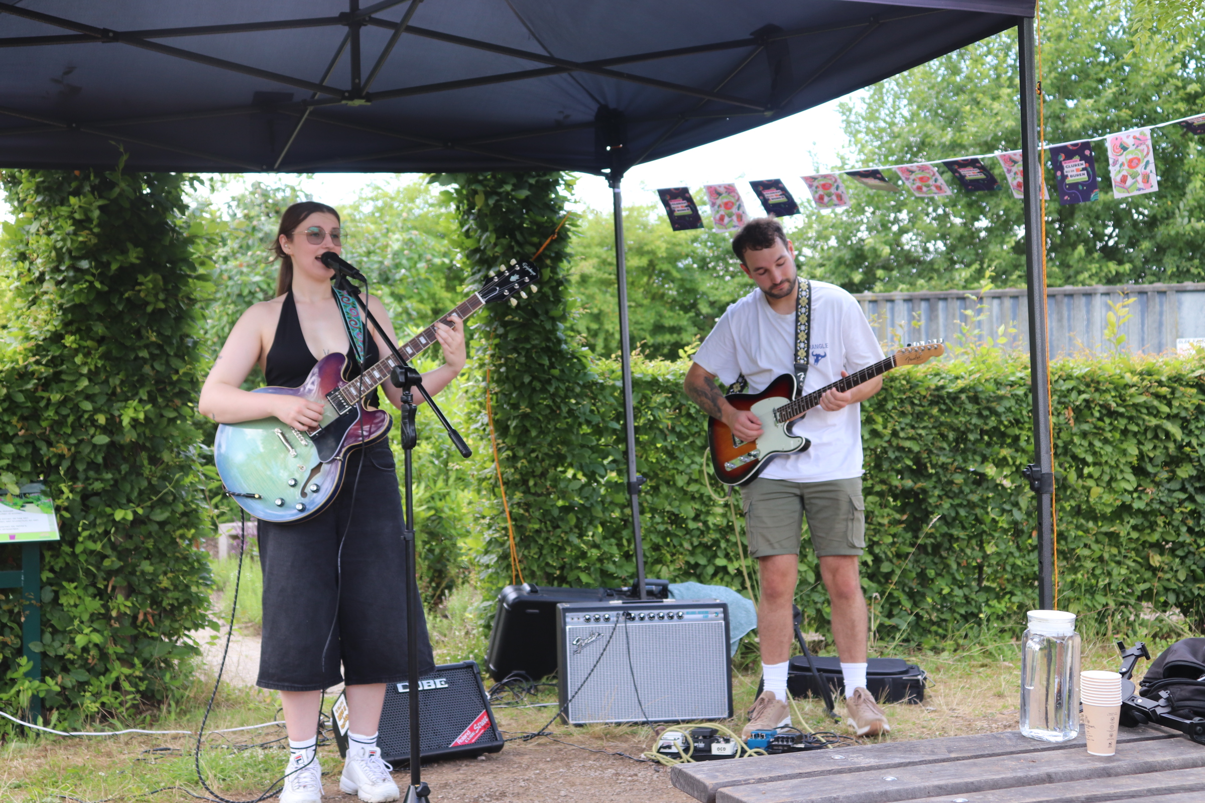 Twee muzikanten spelen gitaar onder een tent in een tuin met groene struiken.