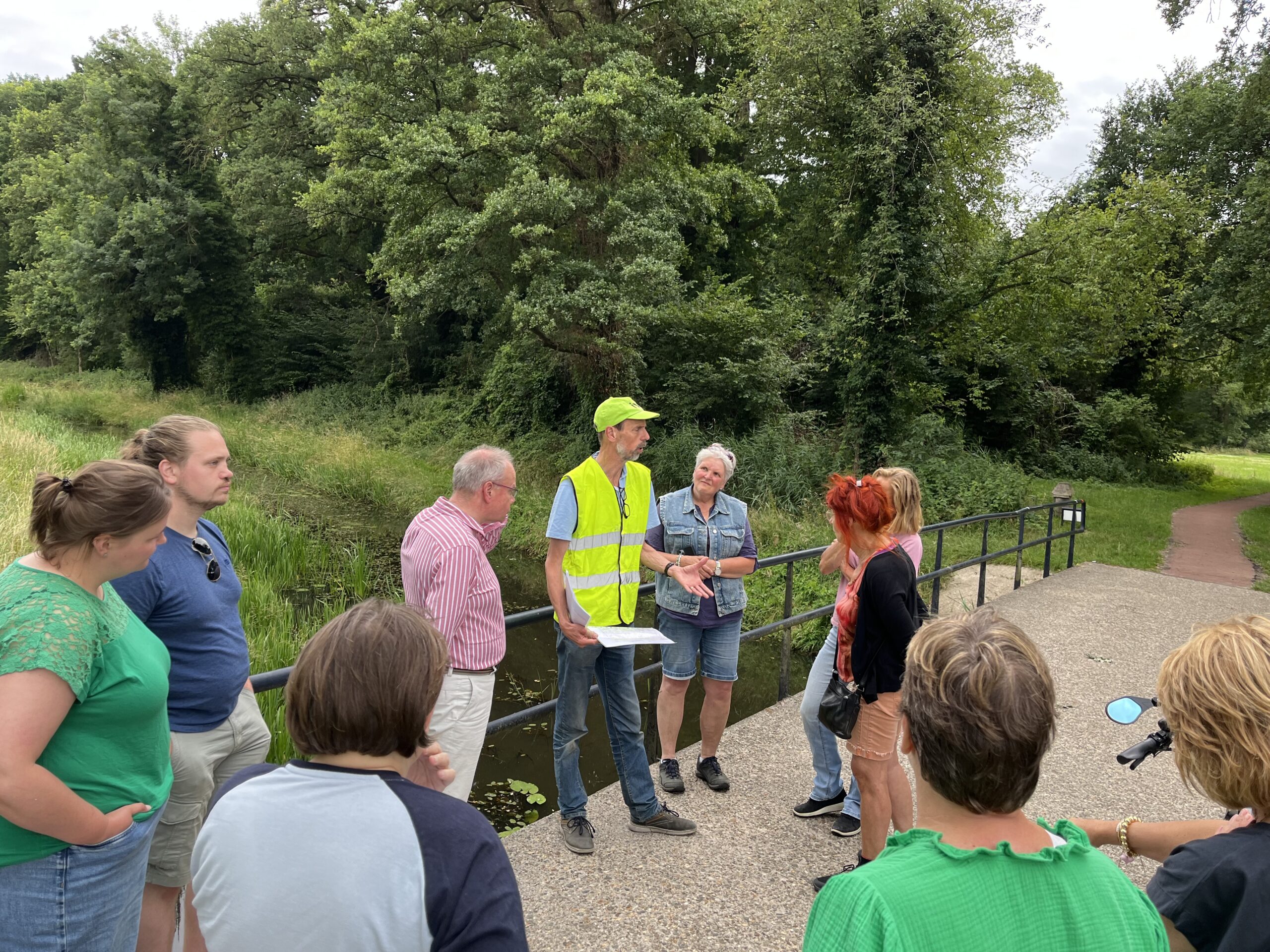 Groep mensen luistert naar een gids in een geel hesje in een groene, bosrijke omgeving.