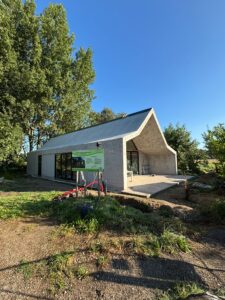 Modern huis met puntdak, grote ramen en terras, omgeven door bomen, onder een heldere blauwe lucht.