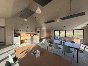 Moderne eetruimte met houten tafels, groene stoelen, hanglampen en grote ramen met uitzicht op grasvelden.