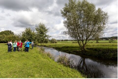 Een groep mensen staat naast een beek in een groen weiland met bomen en een bewolkte hemel.