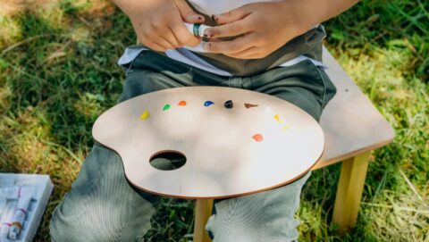 Kind met schilderspalet en verf in het gras, zit op houten krukje.