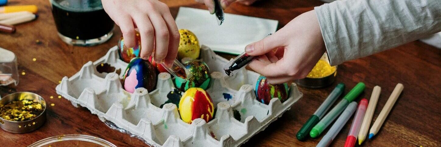 Handen decoreren kleurrijke eieren in een eierdoos, omringd door kunstbenodigdheden.