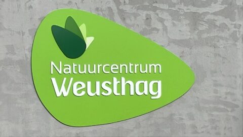 Groen bord met de tekst "Natuurcentrum Weusthag" en gestileerd bladontwerp.