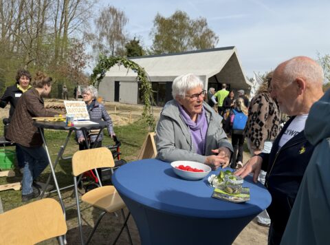 Mensen praten tijdens de opening van een natuurcentrum, met tafels en een modern gebouw op de achtergrond.