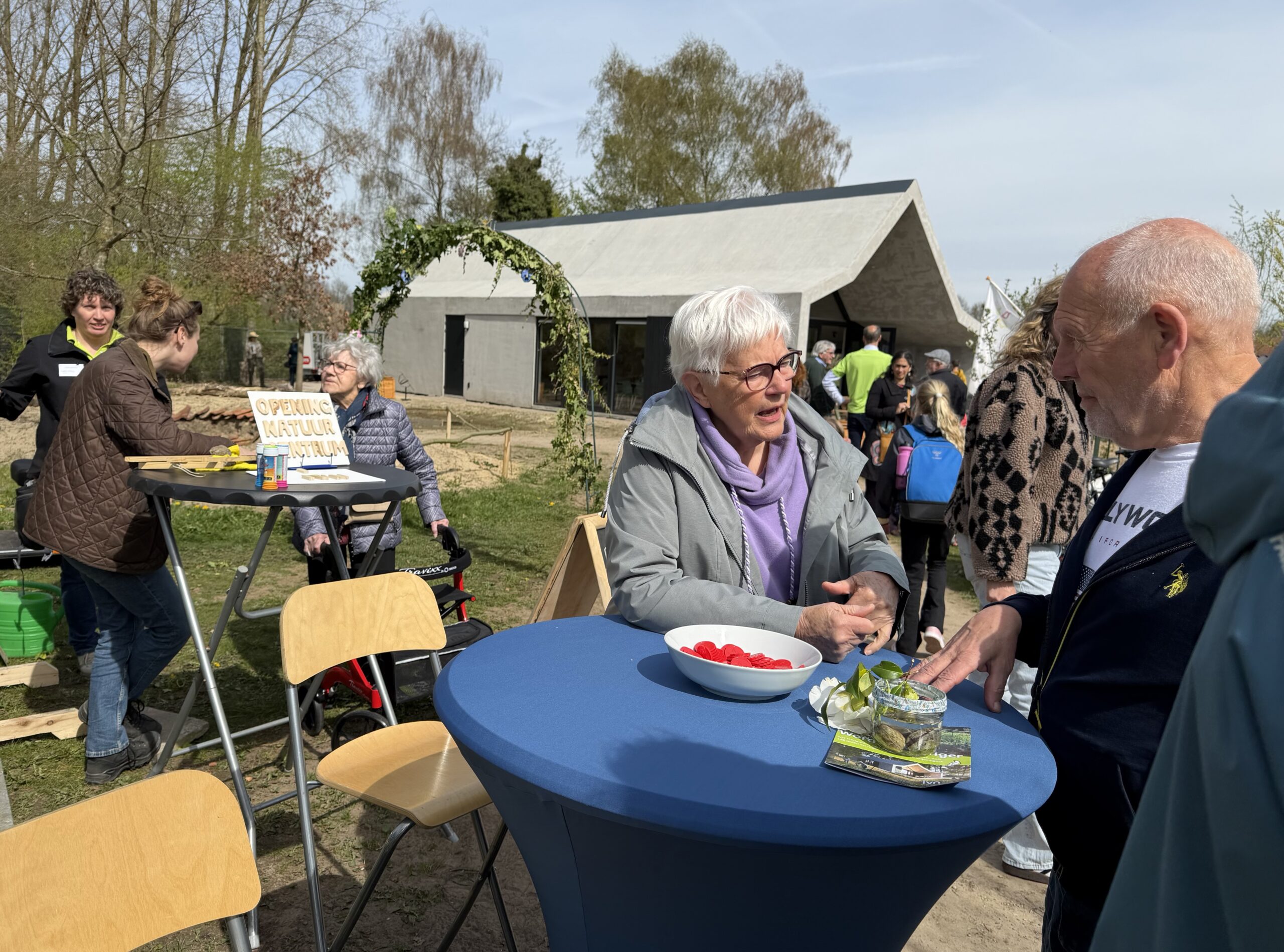 Mensen praten tijdens de opening van een natuurcentrum, met tafels en een modern gebouw op de achtergrond.