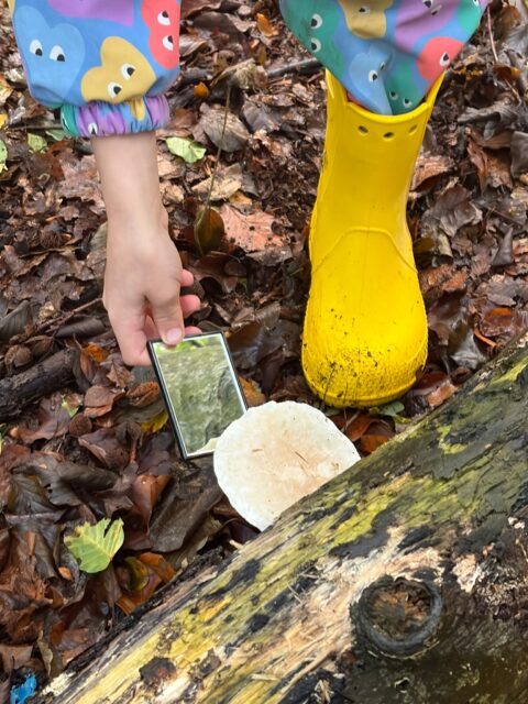 Kind met gele laars inspecteert paddenstoel op bosgrond.