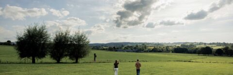 Twee mensen wandelen door een weide met bomen en een wijds, landelijk landschap onder een bewolkte hemel.