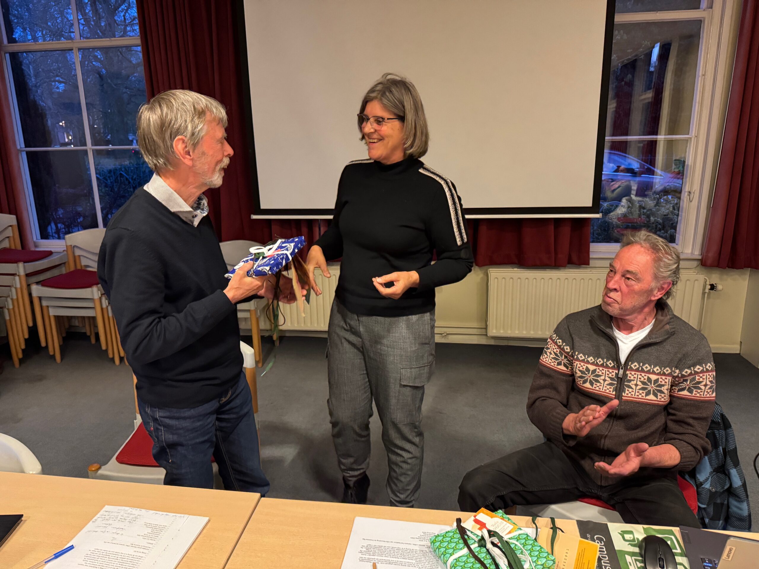 Drie mensen in gesprek, één persoon geeft een geschenk aan een ander in een vergaderruimte.