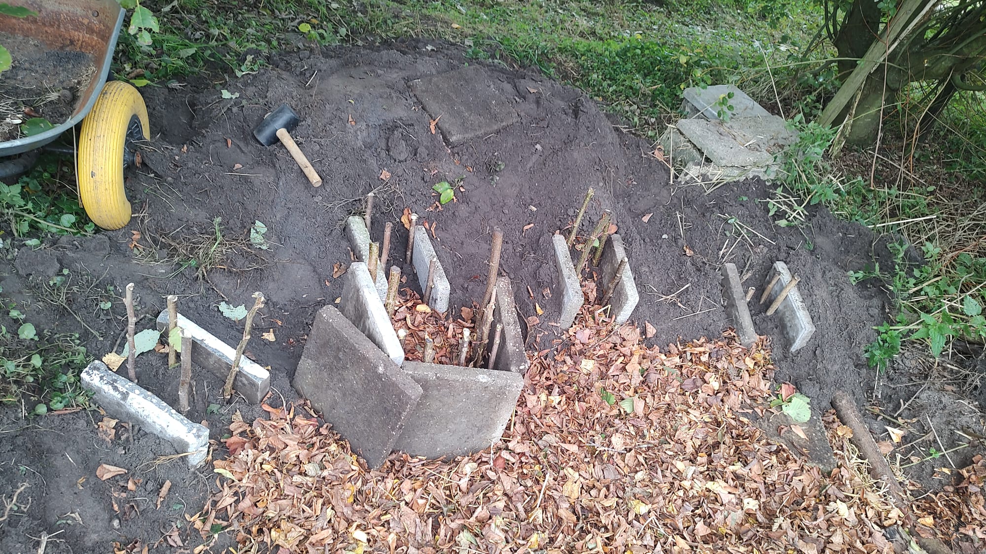 Een constructie van stenen en takken half bedekt met bladeren in een tuin met een kruiwagen.