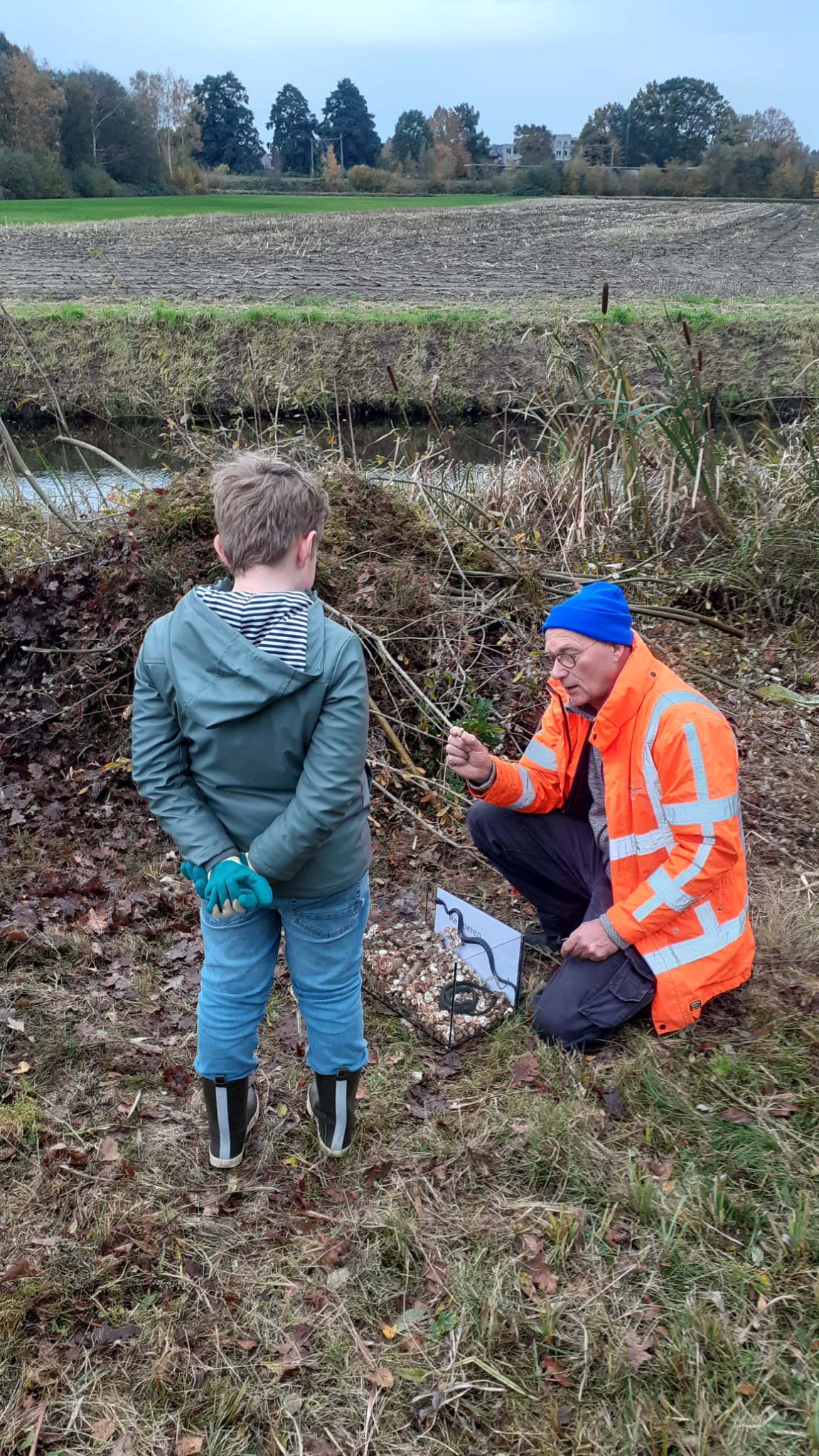 Een man in oranje jas en kind in jas observeren iets in de natuur naast een sloot.