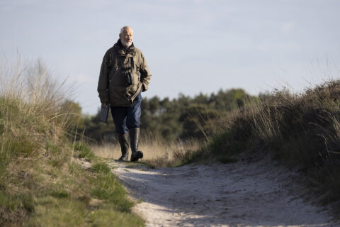 Man wandelt op een zandpad in een natuurgebied, draagt een verrekijker en stevige laarzen.