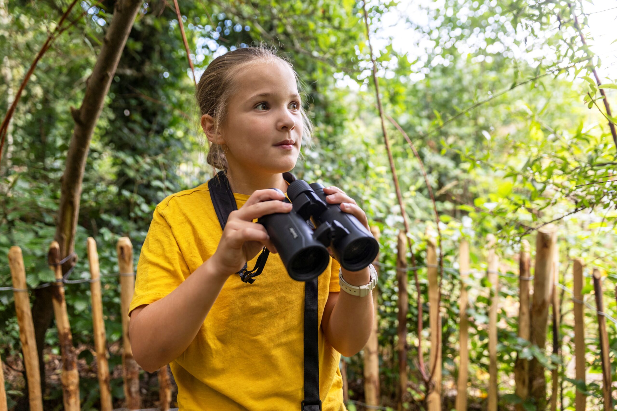 Jong persoon in geel shirt kijkt door verrekijker in een bosrijke omgeving.