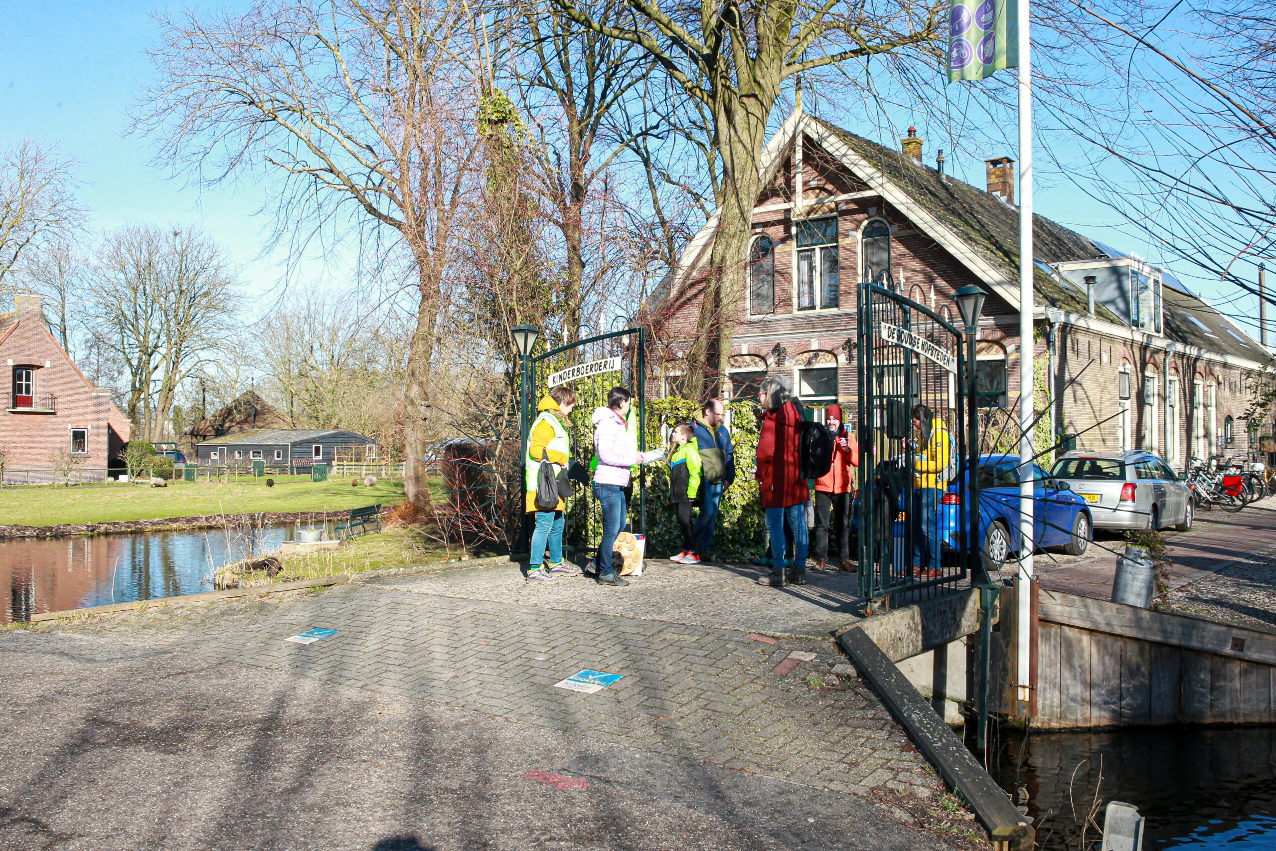 Groep mensen bij de ingang van een kinderboerderij, kleurrijk gekleed, bij een bakstenen huis.