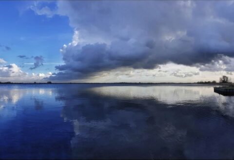 Dreigende wolken boven een kalm meer, met een heldere blauwe lucht aan de linkerzijde.