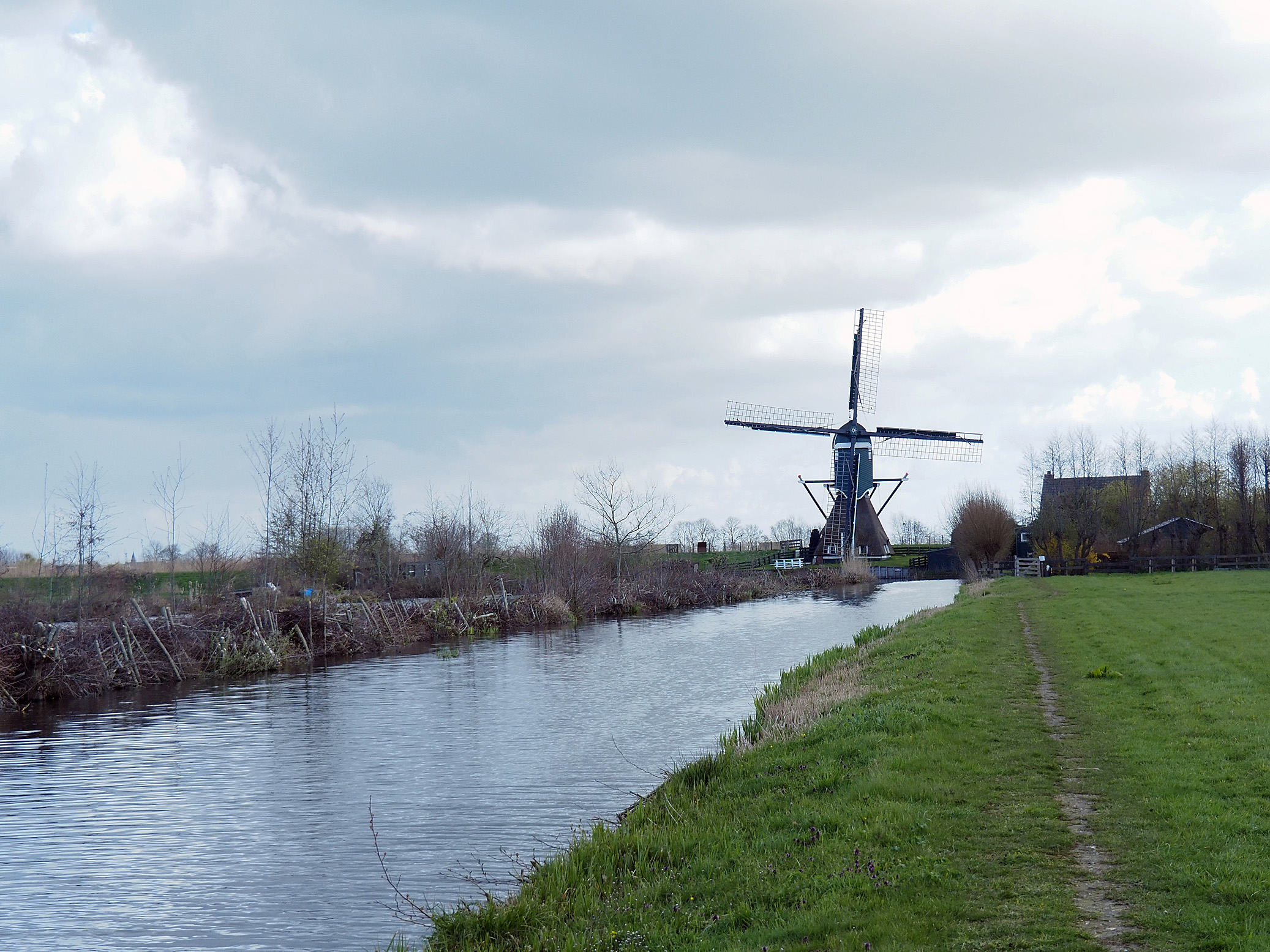 Windmolen naast een kanaal met grasland onder een bewolkte hemel.