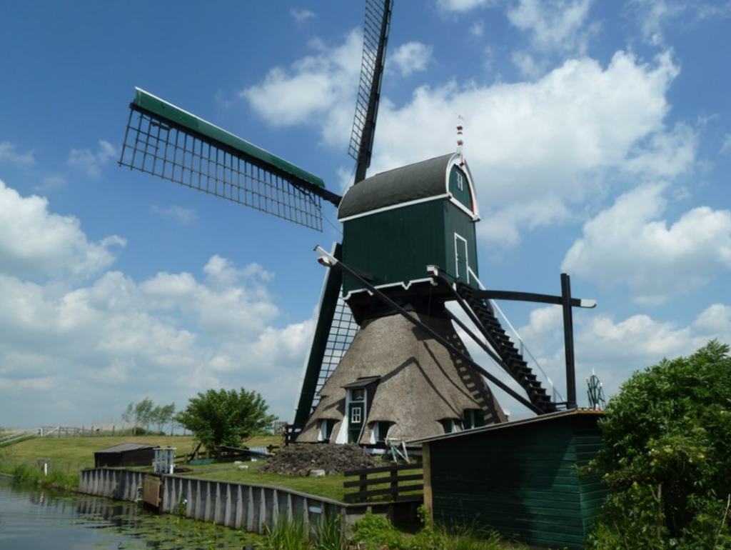 Groene molen met rieten kap naast een sloot, omringd door gras en bomen onder een blauwe lucht.