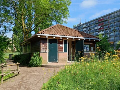 Klein houten gebouw met rode dakpannen, omgeven door bloemen en bomen, naast een breed pad.