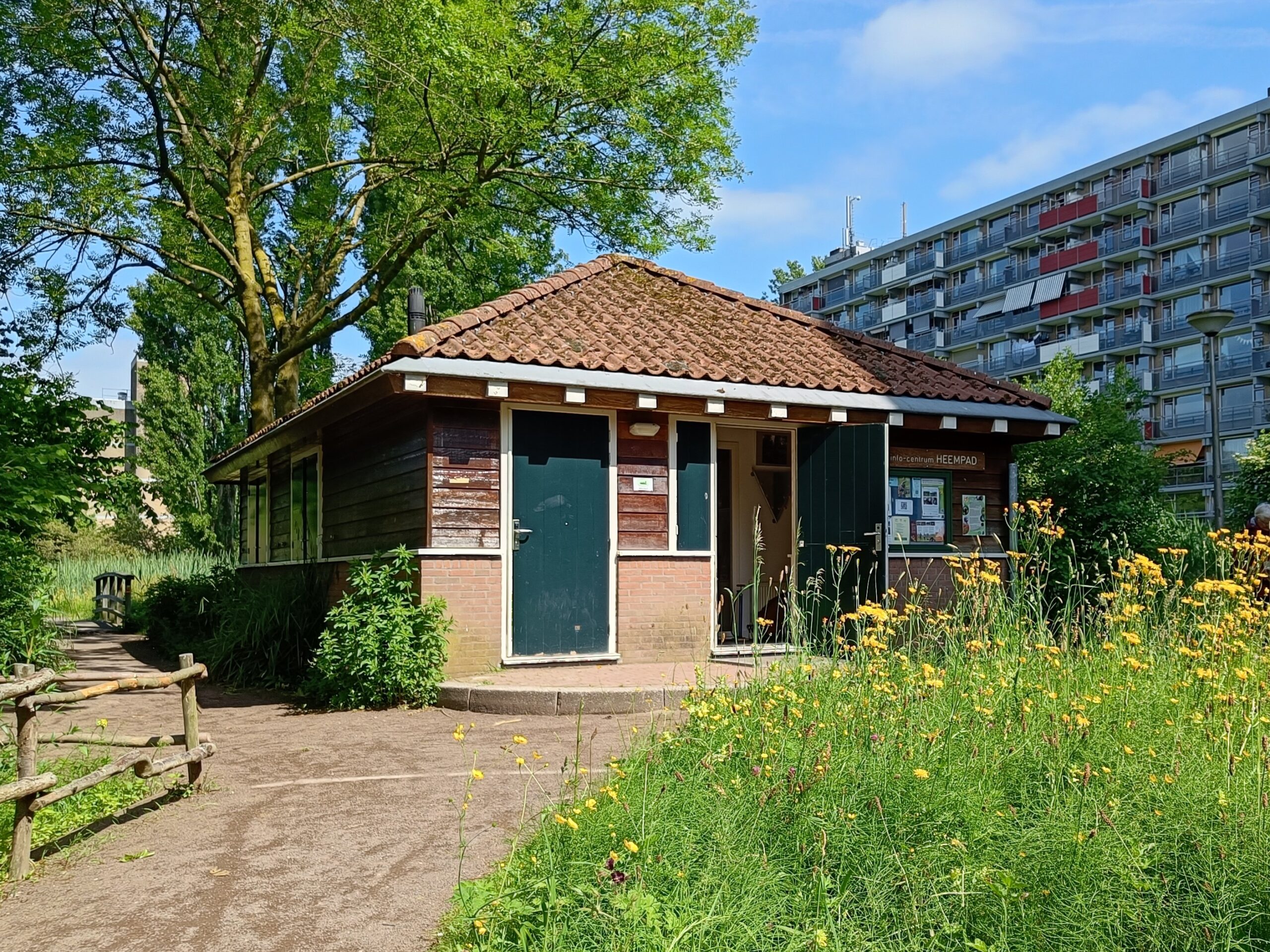 Klein houten gebouw met rode dakpannen, omgeven door bloemen en bomen, naast een breed pad.