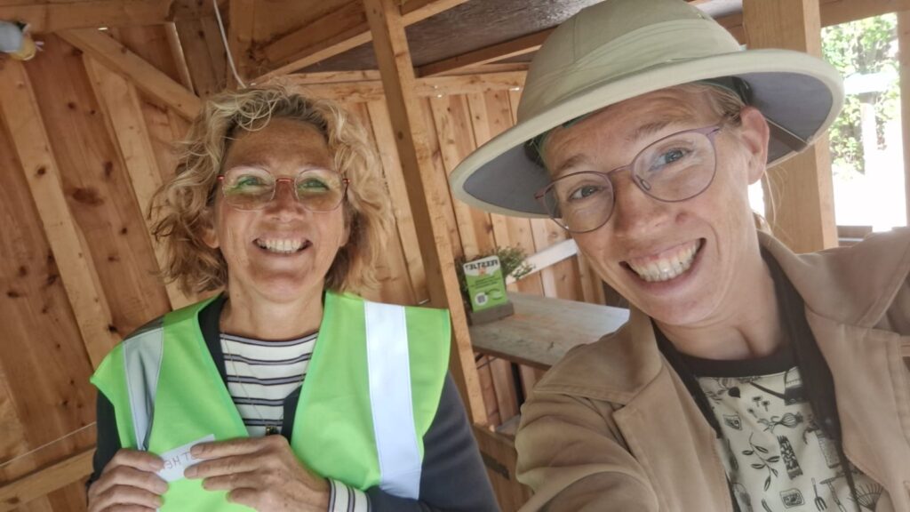 Twee lachende vrouwen in een houten schuur, één draagt een groene veiligheidsvest.