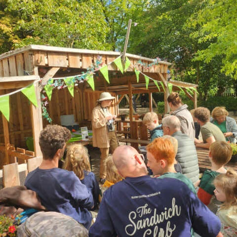 Persoon in safari-outfit geeft presentatie aan een groep buiten, nabij houten schuur met groene vlaggetjes.