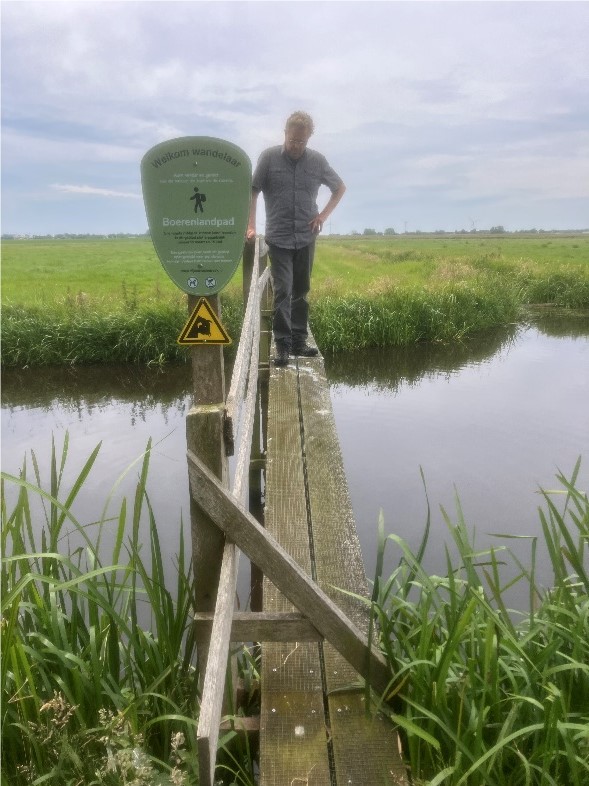 Man staat op smal houten bruggetje over een sloot, met een bord "Boerenlandpad" naast hem.