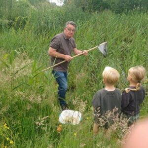 Man met schepnet in gras bij kinderen, emmer op de grond in natuuromgeving.