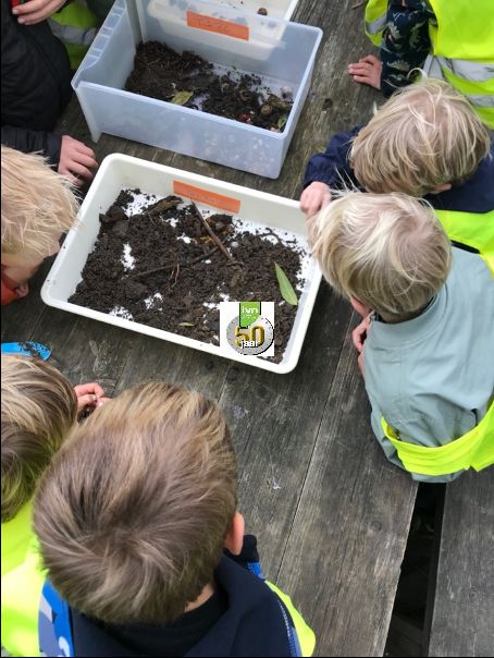 Groep kinderen onderzoekt bakken met aarde en bladeren op een houten tafel buiten.