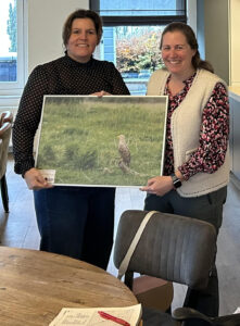 Twee mensen tonen samen een grote foto van een vogel in een grasveld, in een woonkamer.